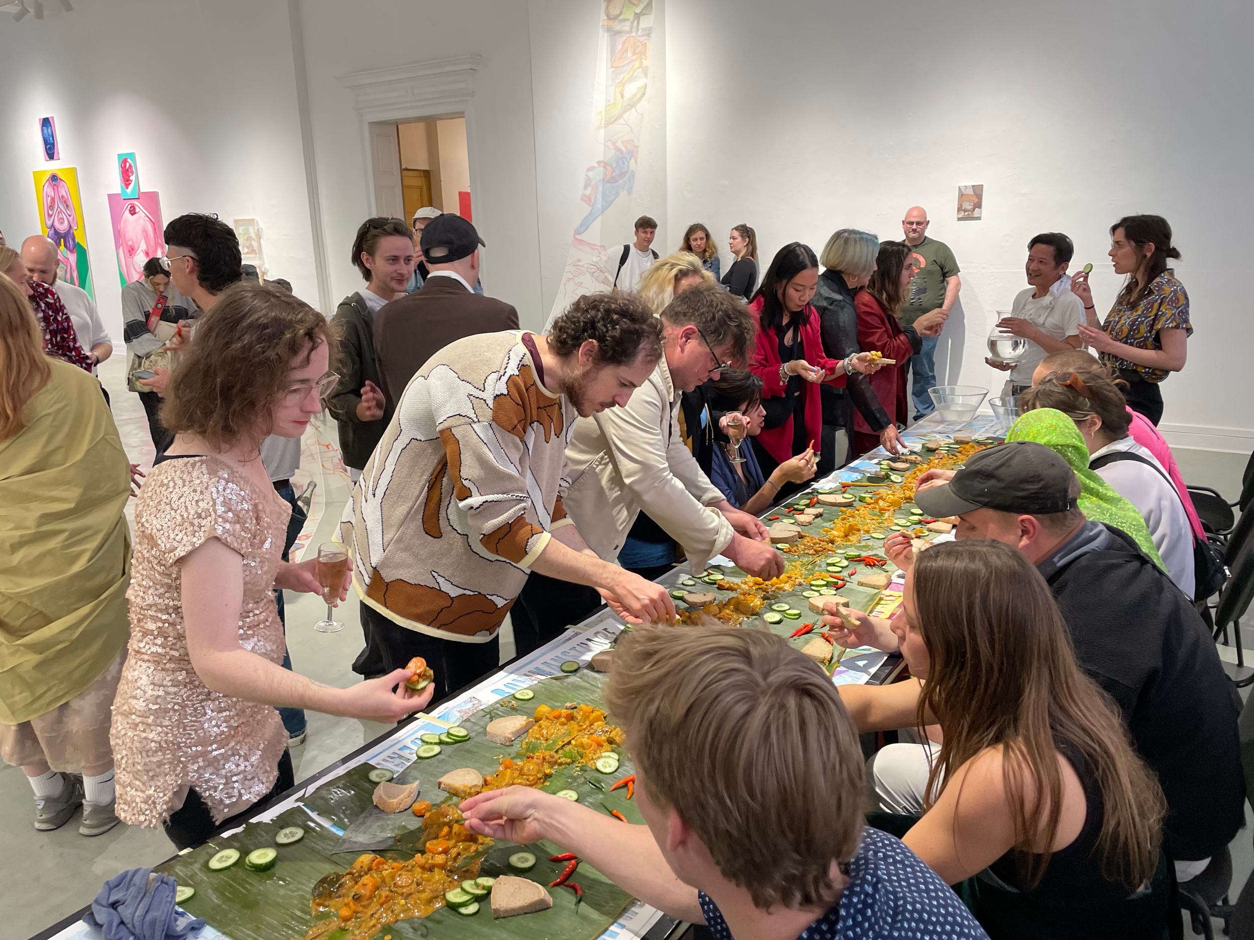 People gathered around a long table with food at an art gallery, viewing and serving meals, with artwork on the white walls in the background.