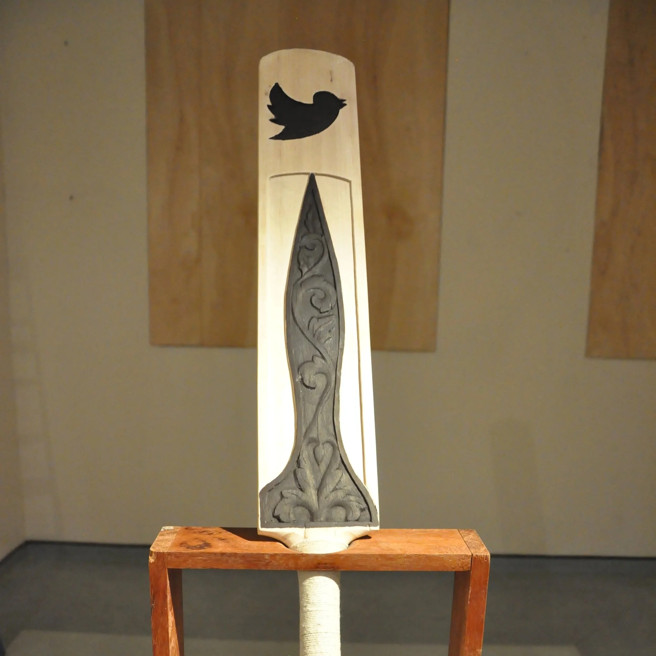A wooden sculpture featuring a black silhouette of a bird perched on a branch, mounted on a stand with a wooden base.