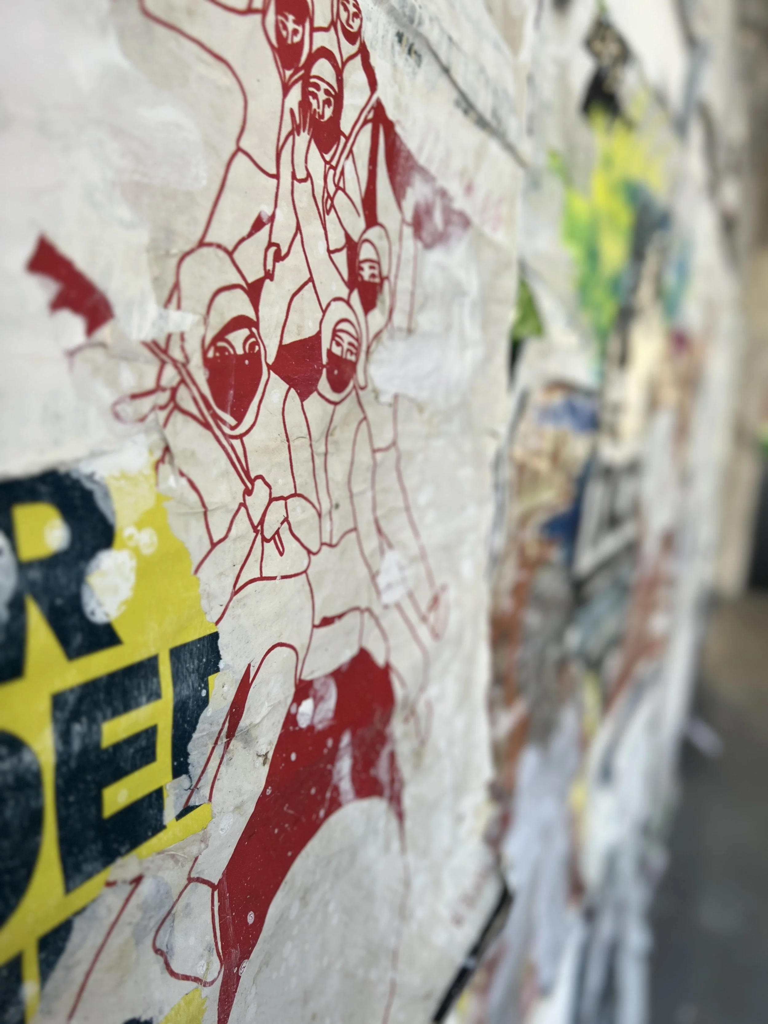 Close-up of a torn and wrinkled paper poster featuring line art illustrations of soldiers and military equipment in red ink, on a textured surface with other colorful posters in the background.