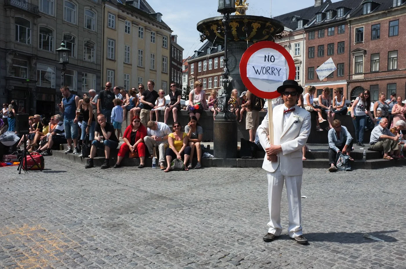 A man in a white suit and black hat holding a sign that says 'NO WORRY' standing in front of a crowd sitting on steps around a fountain in an urban square with historic buildings.