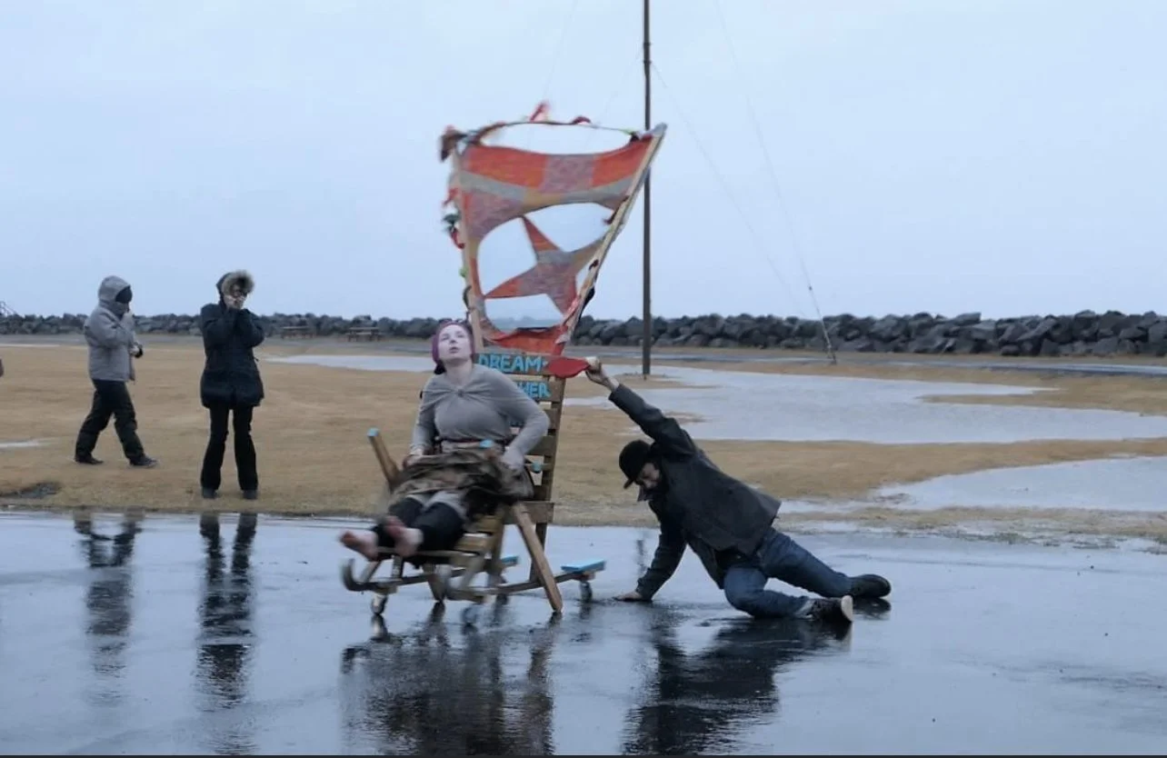 People near a seaside with a broken wind sock, one person sitting on a deck chair, another person pushing the chair, and two others standing in the background.