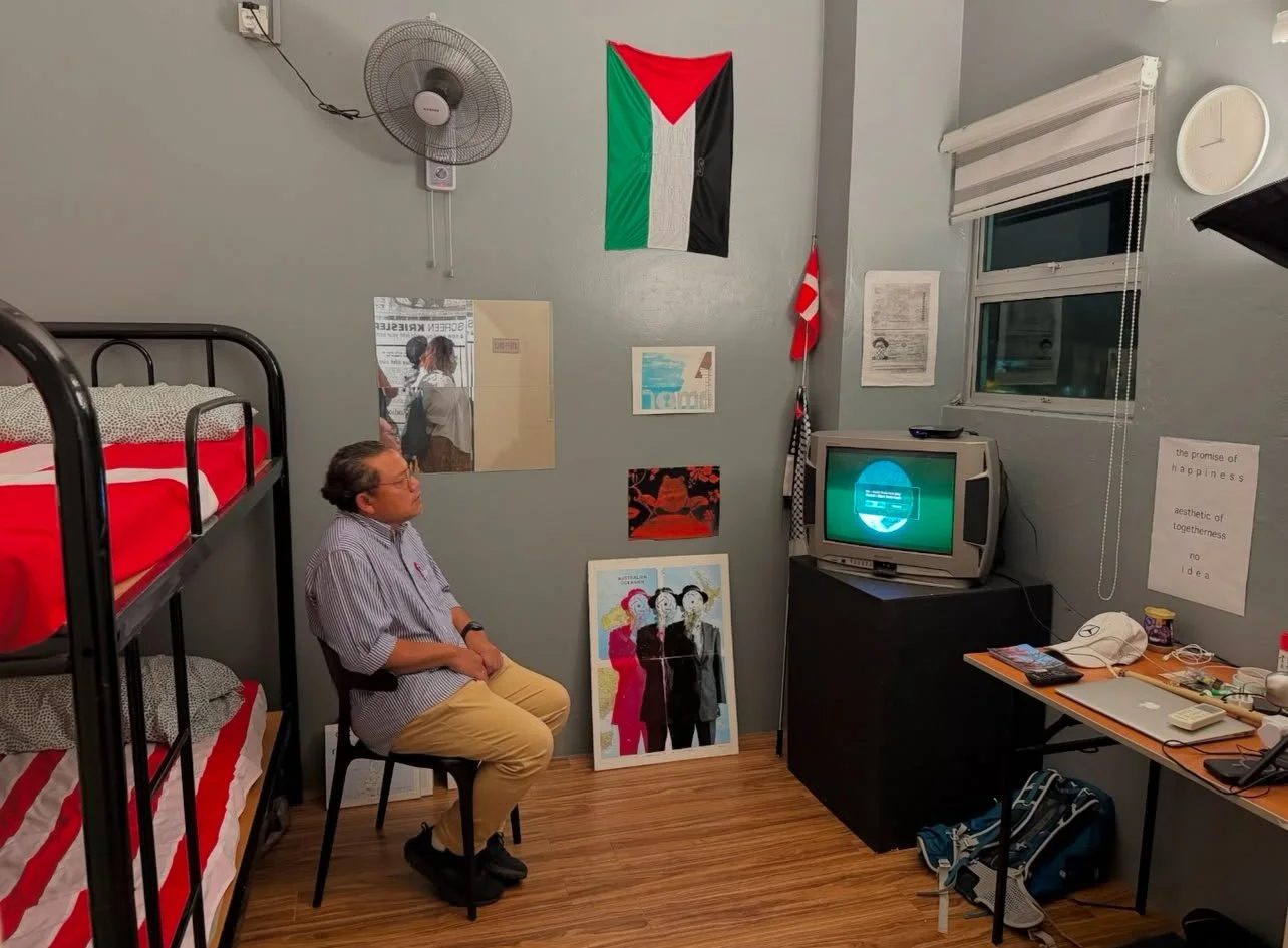 A person sitting on a chair in a small, decorated room with a bunk bed, wall art, a television, and a desk with various items. The room has a Palestinian flag and a Danish flag on the wall, a fan, and a clock.