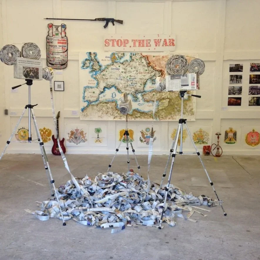 Installation art featuring a pile of rolled-up newspapers or paper strips on the floor with four tripods holding projectors aimed at a backdrop that includes a map of Europe and surrounding regions, a sign saying "Stop the War," and various flags, artwork, and photographs on the walls.