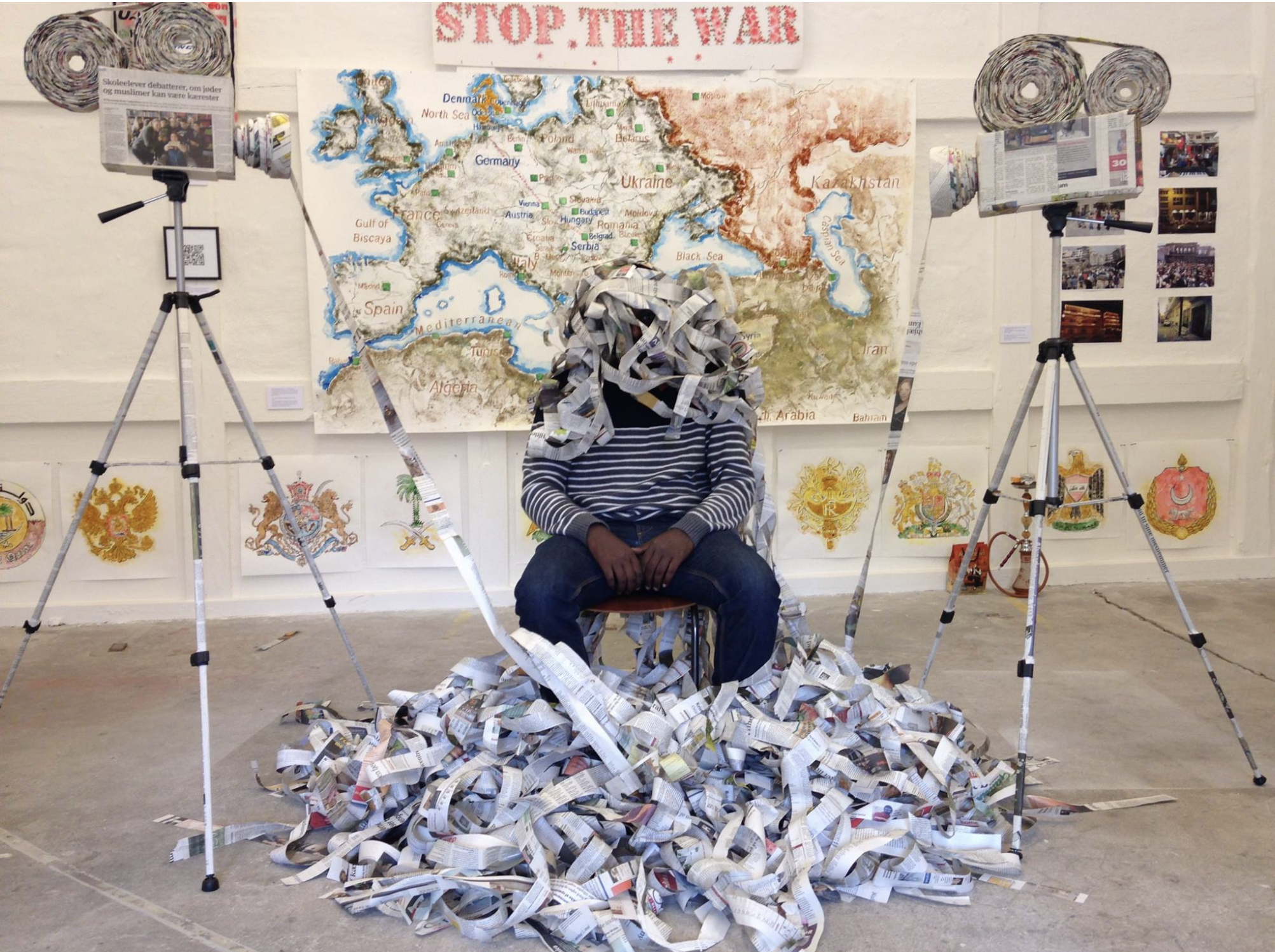 Person sitting on a chair, covered in shredded newspaper, in front of a large map of Europe and parts of Asia and Africa. There are two tripods with rolled newspaper on either side of the person, and the floor is scattered with shredded newspaper.