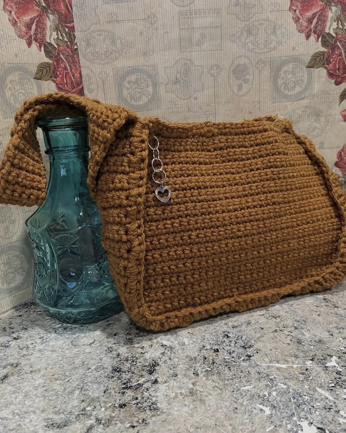 A clear blue glass bottle, placed next to a brown crochet handbag with a chain and heart charm, on a granite countertop against a floral and vintage wallpaper background.