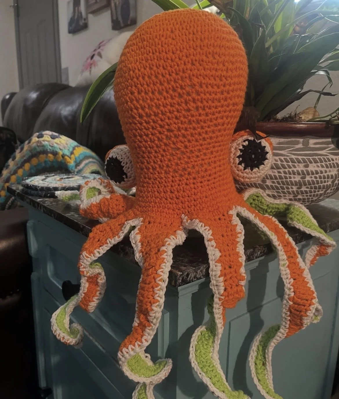 A large, orange, crocheted octopus plush toy with black and white eyes, placed on a kitchen counter next to plants and other crocheted items.