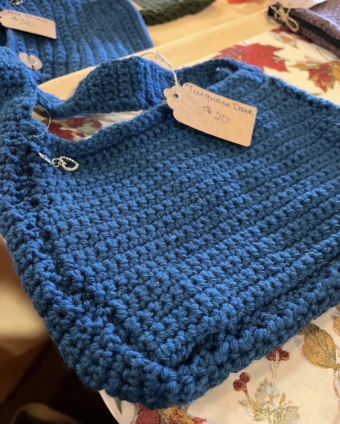 A blue crocheted purse with a textured pattern is placed on a table with decorative autumn leaves. There is a handwritten tag attached to it that reads 'Turquoise Down $30'.