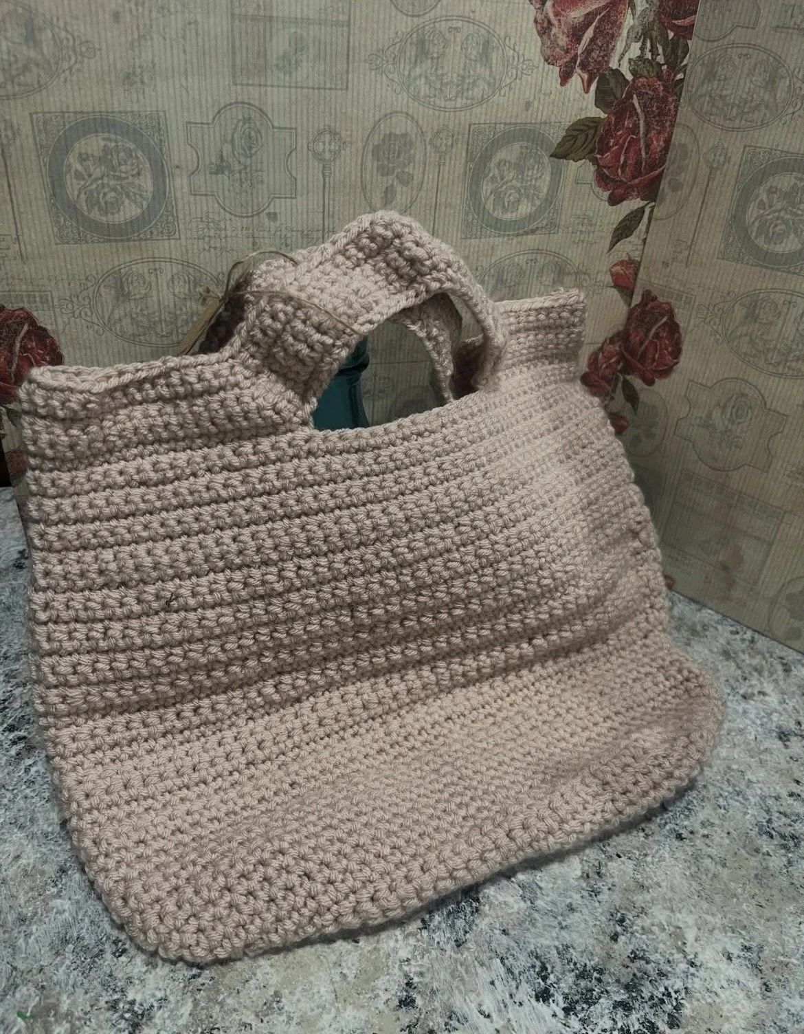A hand-knit beige tote bag with two handles is placed on a textured surface against a floral and geometric patterned background.