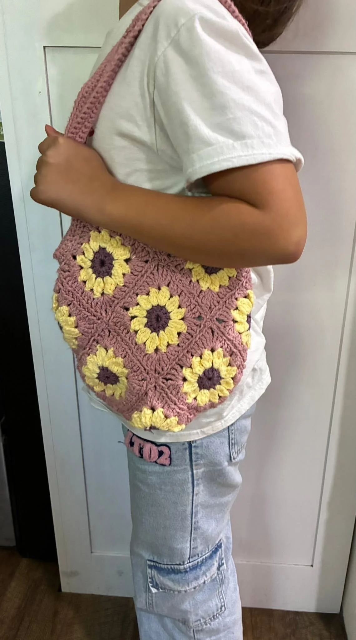 Child wearing a white t-shirt, light blue jeans with patches, and carrying a pink crocheted bag with yellow and purple flower patterns.
