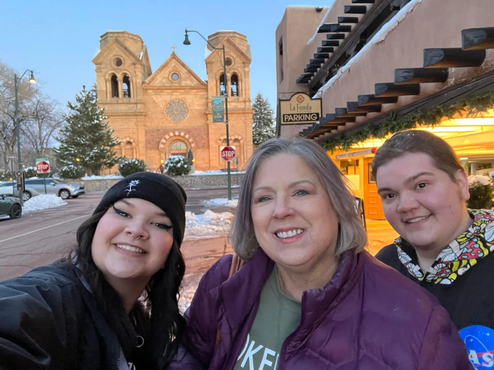 Three smiling people, two women and one man, taking a selfie outdoors in winter, with a historic church in the background illuminated at dusk, snow on the ground, and a building with a sign that reads "La Fonda" nearby.