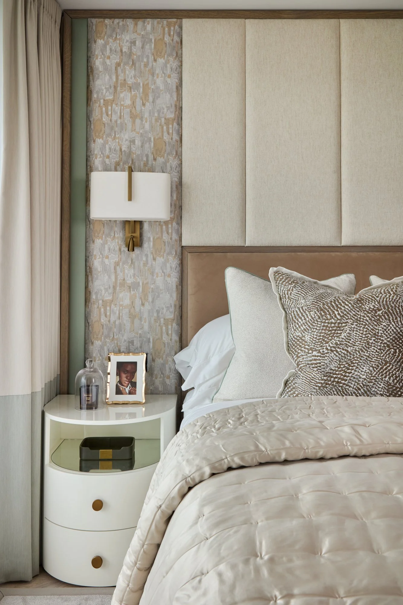Contemporary bedroom with a full-height, full-width bespoke upholstered headboard, decorative cushions, patterned wallpaper, wall-mounted bedside light and a white circular side table