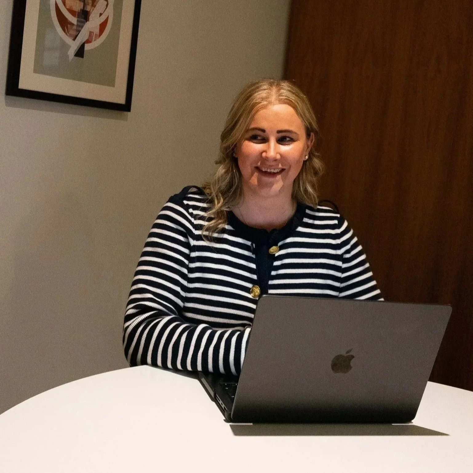 A woman with blonde hair wearing a black and white striped long sleeve shirt, sitting at a table with an open MacBook laptop in front of her. She is smiling and looking to her right in a room with a framed piece of abstract artwork on the wall behind her.