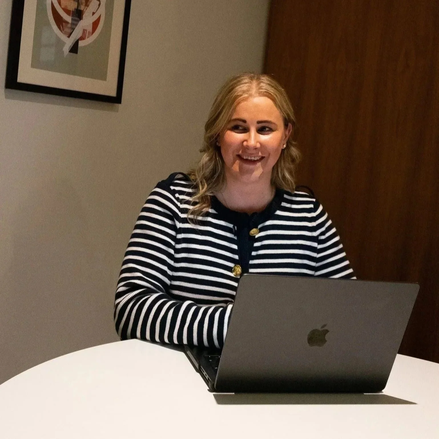 A woman with blonde hair sitting at a white table, smiling, working on a silver laptop with an Apple logo. She is wearing a black and white striped cardigan with gold buttons and a black top underneath.