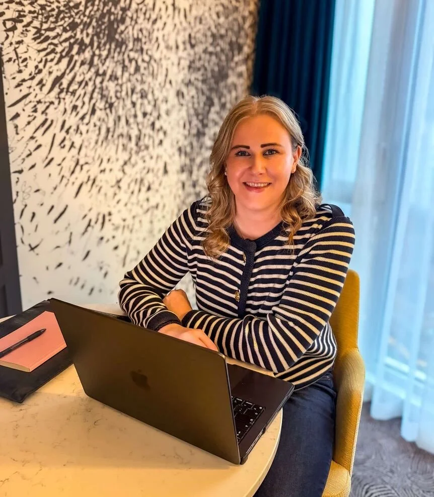 A woman with blonde hair sitting at a round table with a laptop, a pink notebook, and a pen, smiling at the camera in a well-lit room with patterned wallpaper and blue curtains.