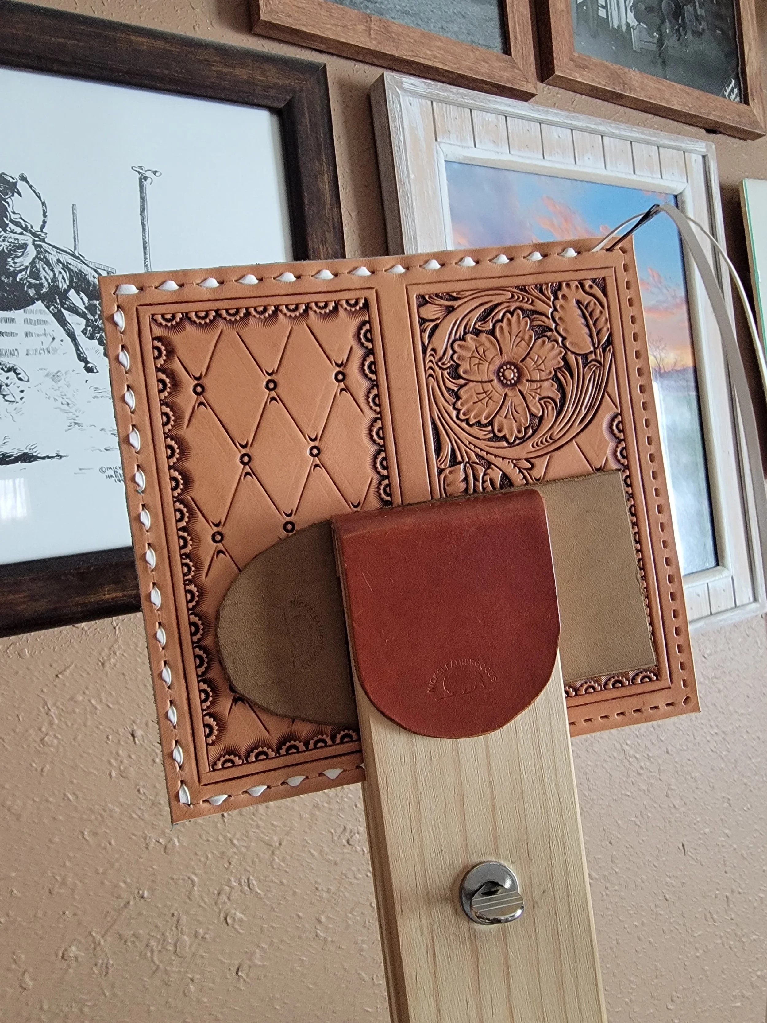 Close-up of tooled leather work on stitching pony in front of a decorative leather panel with floral and diamond patterns, against a wall with framed artwork.