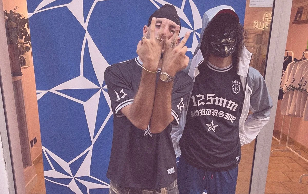 Two men are posing for a photo inside a store. One man on the left is making a hand gesture and is wearing a black t-shirt with white lettering, along with rings, a watch, and a bracelet. The man on the right is wearing a black mask, a hooded jacket, and a black and white sports shirt with white lettering and graphics. They are standing in front of a geometric blue and white patterned backdrop, with shelves displaying clothing and accessories visible on the sides.