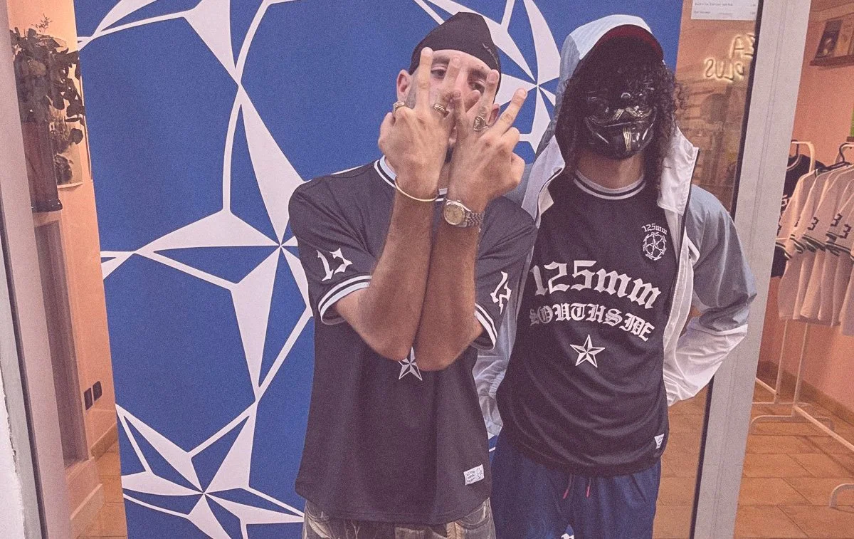 Two young men standing in front of a blue geometric mural inside a clothing store. One is wearing a black beanie and a black sports jersey, making hand gestures, wearing rings, and a watch. The other is wearing a hooded jacket over a black t-shirt with white text and graphics, including a star, and a black mask with a design.