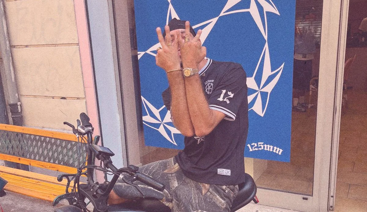 A person sitting on a motorbike with their face covered by their hands, standing in front of an 'NATO' sign with geometric star patterns.
