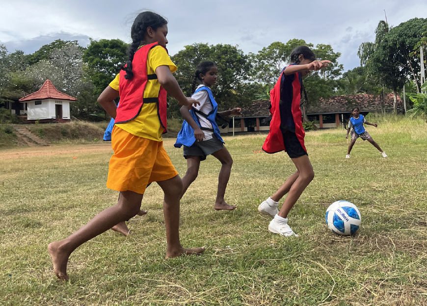 Vier Kinder spielen Fußball auf einem offenen Feld, eines schießt nach vorne, während die anderen zuschauen. Im Hintergrund Bäume und Gebäude, das Wetter ist bewölkt.
