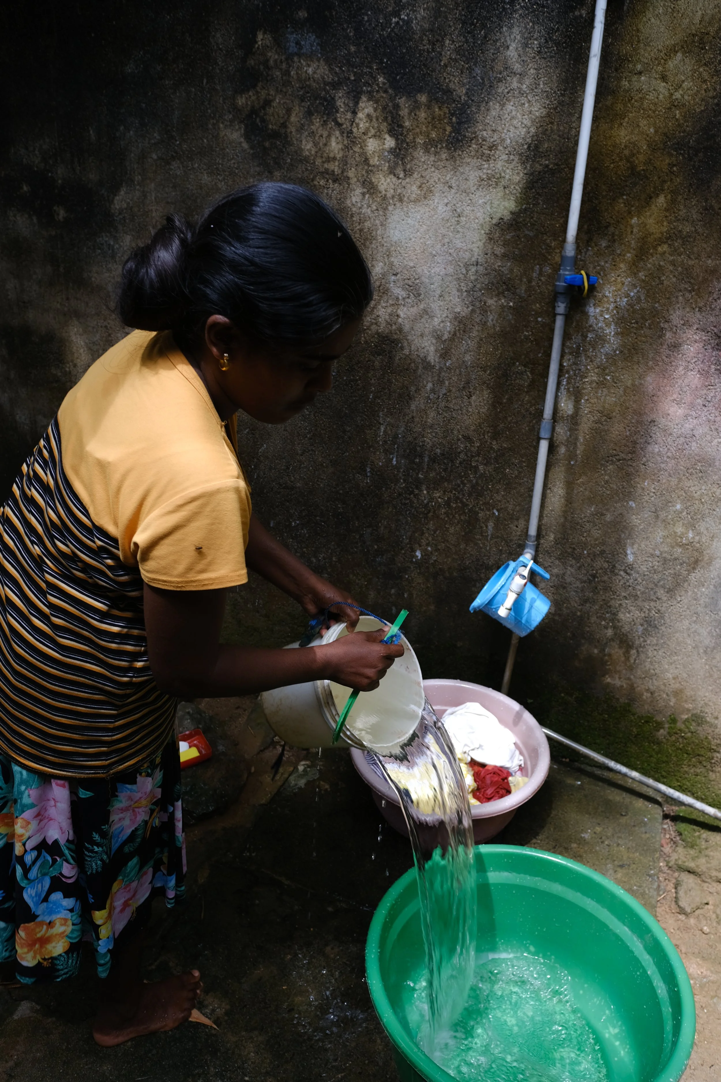 Frau beim Wäschewaschen in einer grünen Tonne, Wasser läuft hinein, an der Wand ist ein Wasserrohr mit Wasserhahn, der auf eine blaue Wasserwaage geklemmt ist.