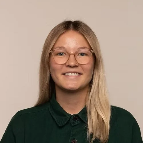 Eine junge Frau mit langen blondem Haar, Brille und grünem Hemd lächelt vor einem neutralen Hintergrund.