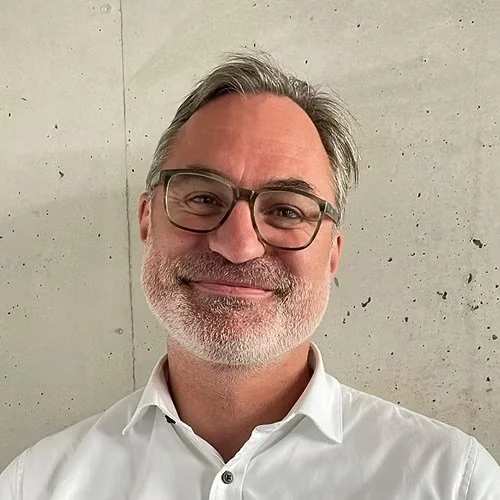 Porträt eines lächelnden Mannes mit Brille vor einer Betonwand, trägt ein weißes Hemd.