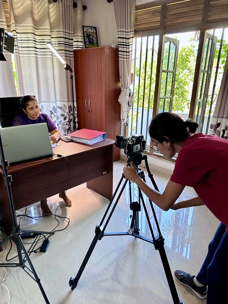 Aufnahme eines Video- oder Foto-Setups in einem Büro mit einer Frau, die vor einem Schreibtisch sitzt, und einem Fotografen, der eine Kamera auf einem Stativ benutzt.