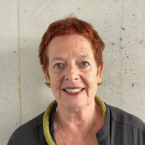 Nahaufnahme einer älteren Frau mit kurzen roten Haaren, die lächelnd in die Kamera schaut, vor einer Betondecke.