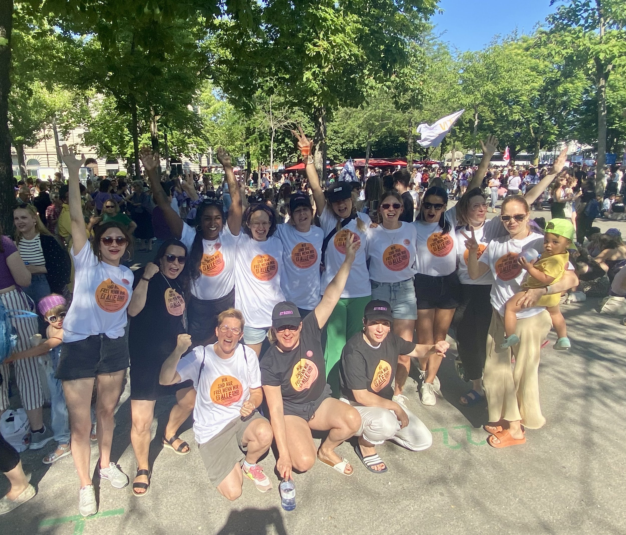 Gruppe von Menschen bei einer Demonstration im Freien, viele tragen T-Shirts mit Slogans, einige machen Peace-Zeichen, im Hintergrund sind viele weitere Menschen und Bäume zu sehen.
