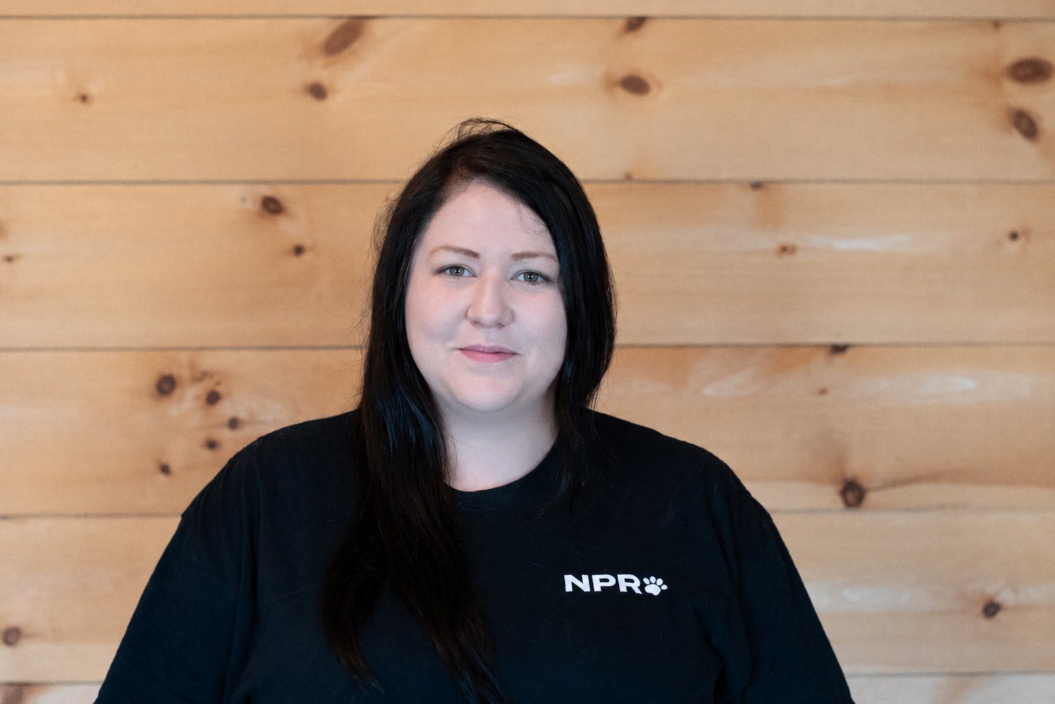 A woman with long dark hair, wearing a black NPR t-shirt, standing in front of a wooden paneled wall.