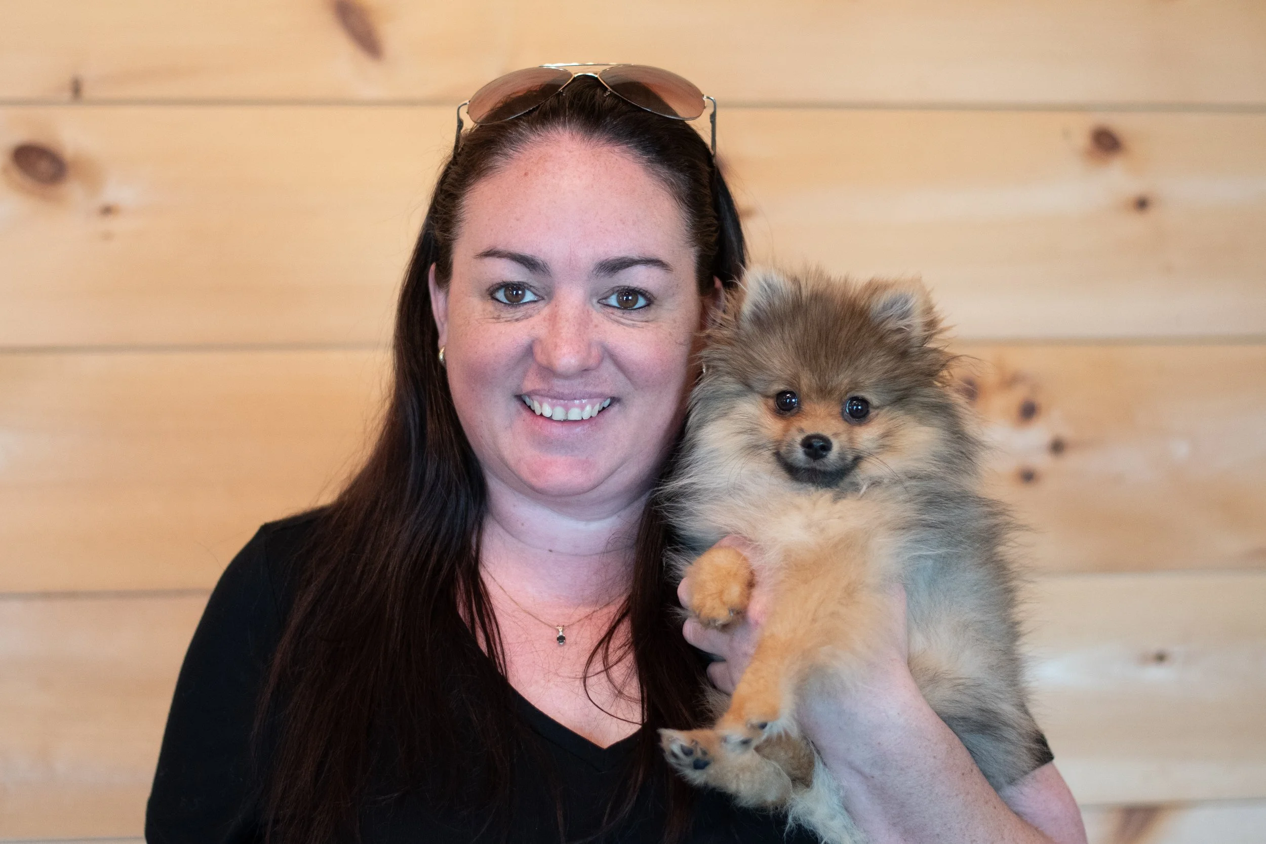A woman with long brown hair, wearing sunglasses on her head, smiling while holding a fluffy puppy with brown, black, and gray fur in front of a wooden background.