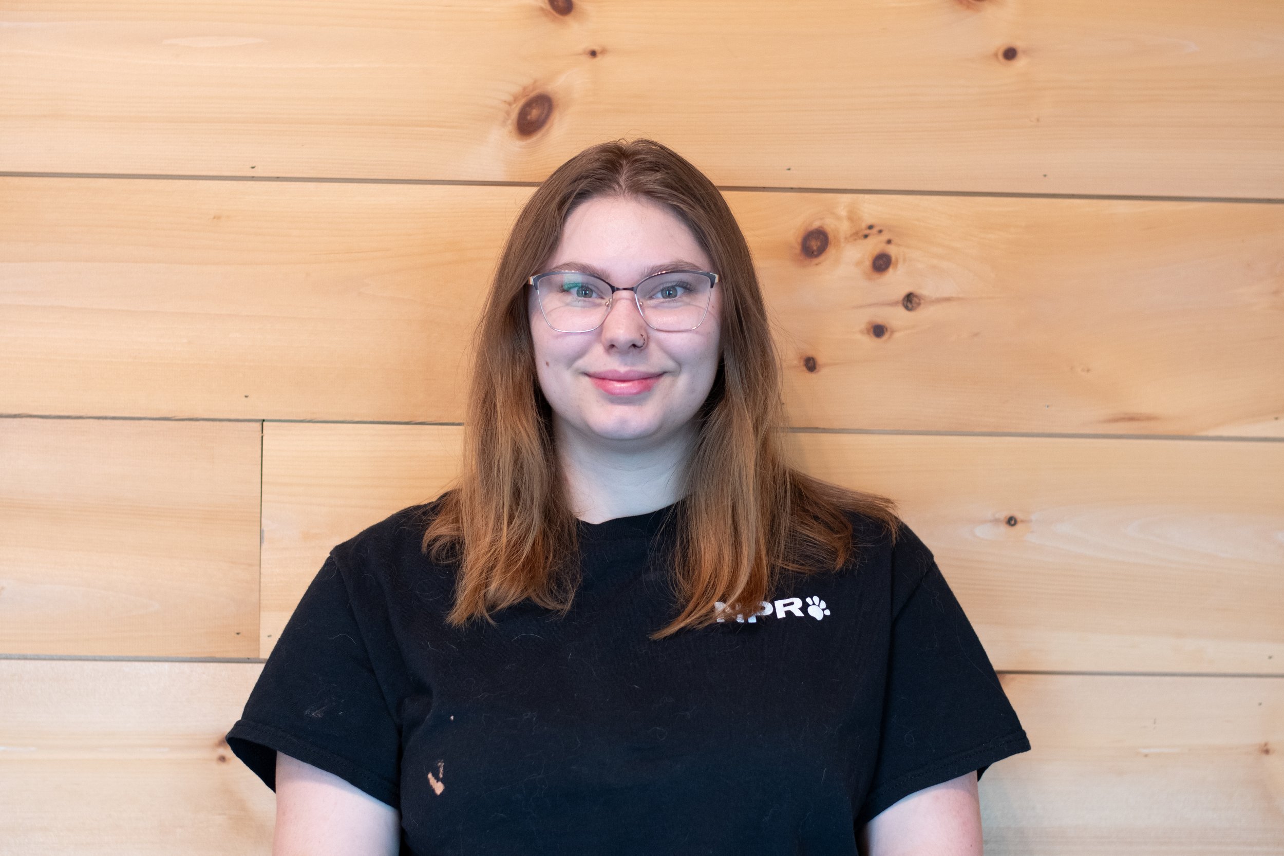 A young woman with long, red hair and glasses sitting in front of a wood-paneled wall, wearing a black T-shirt with partial white text.