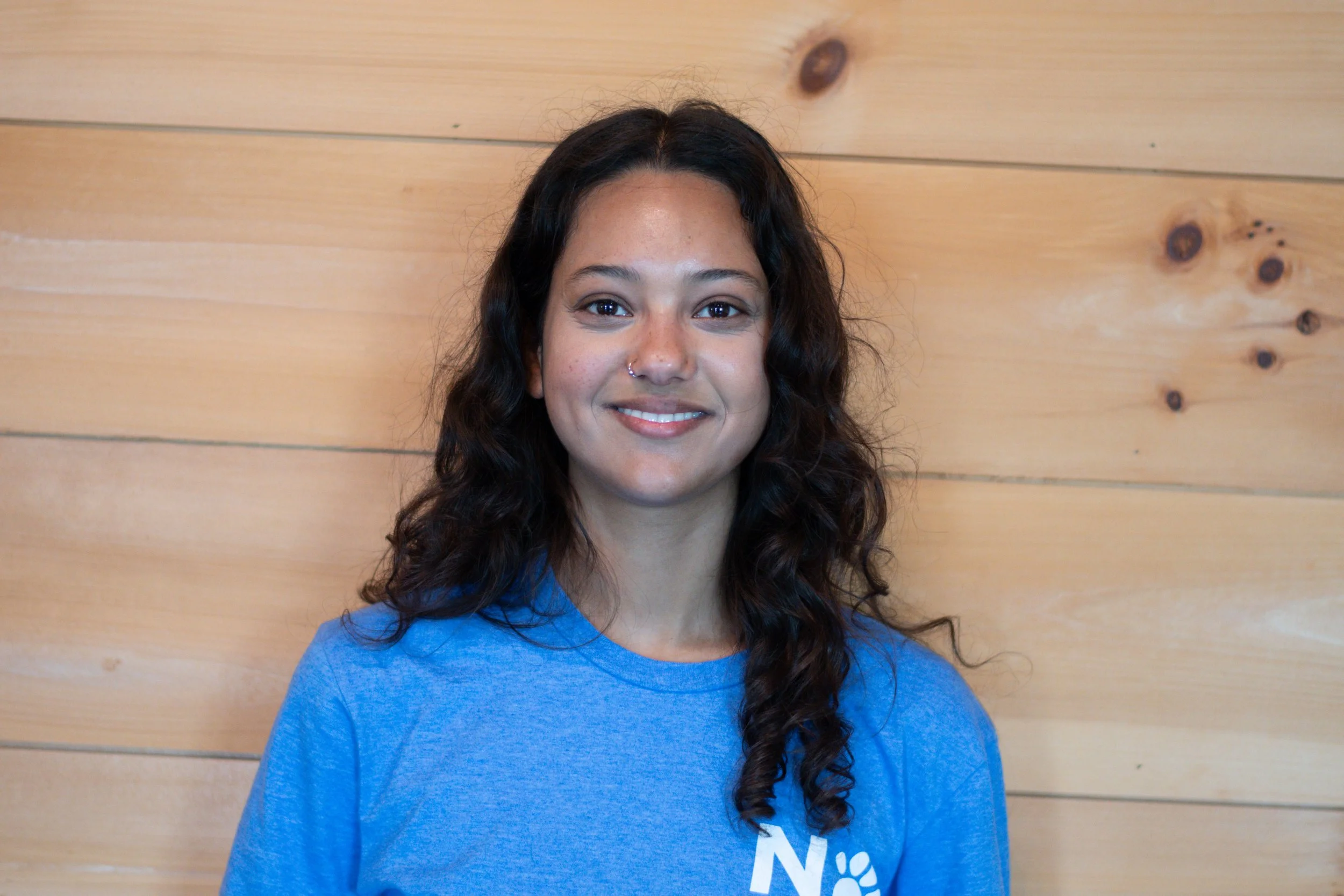 A young woman with dark, curly hair and a nose piercing wearing a blue T-shirt stands in front of a wooden wall.