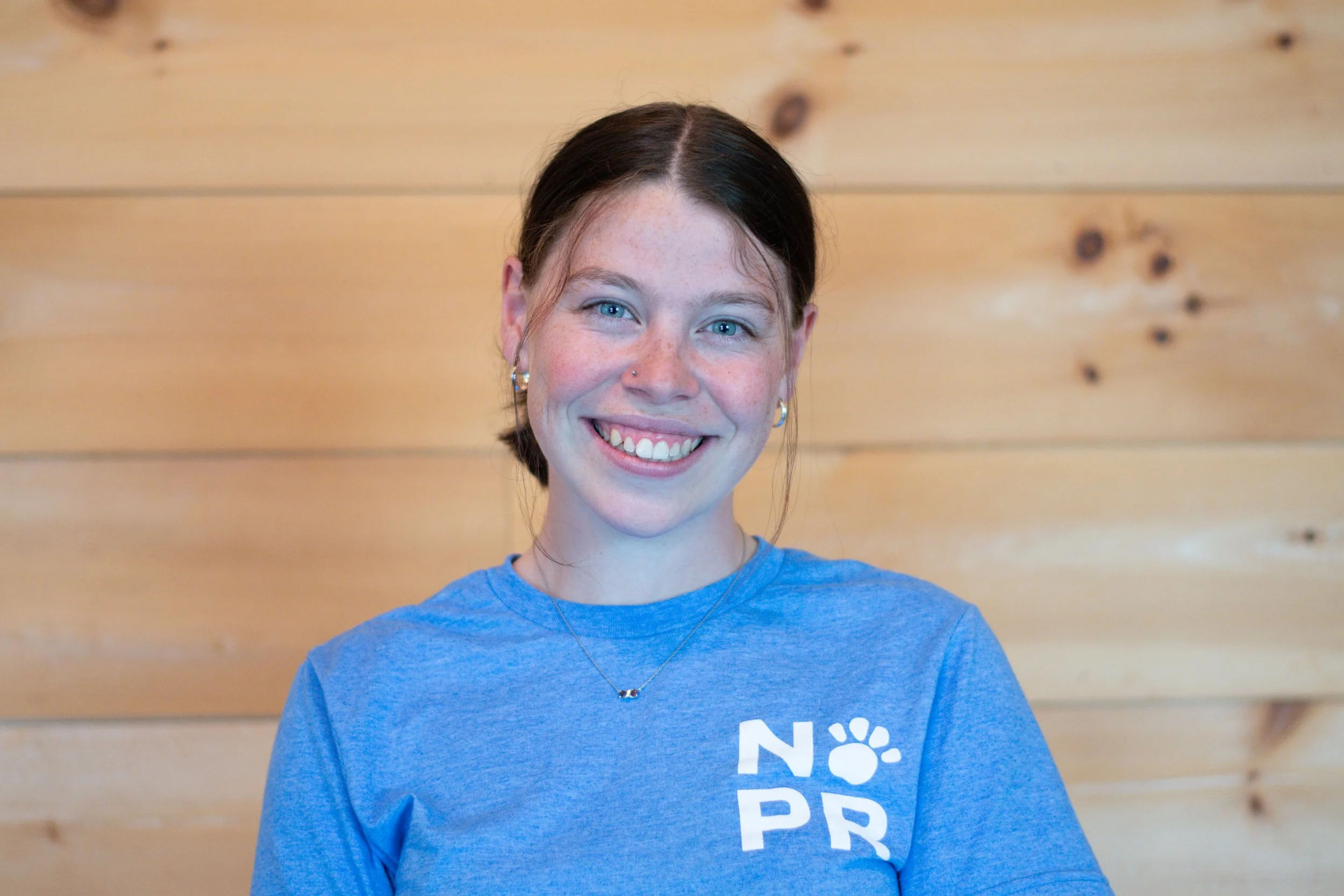 A young woman with blue eyes, dark brown hair tied back, wearing a blue T-shirt and silver jewelry, smiling in front of a wooden background.