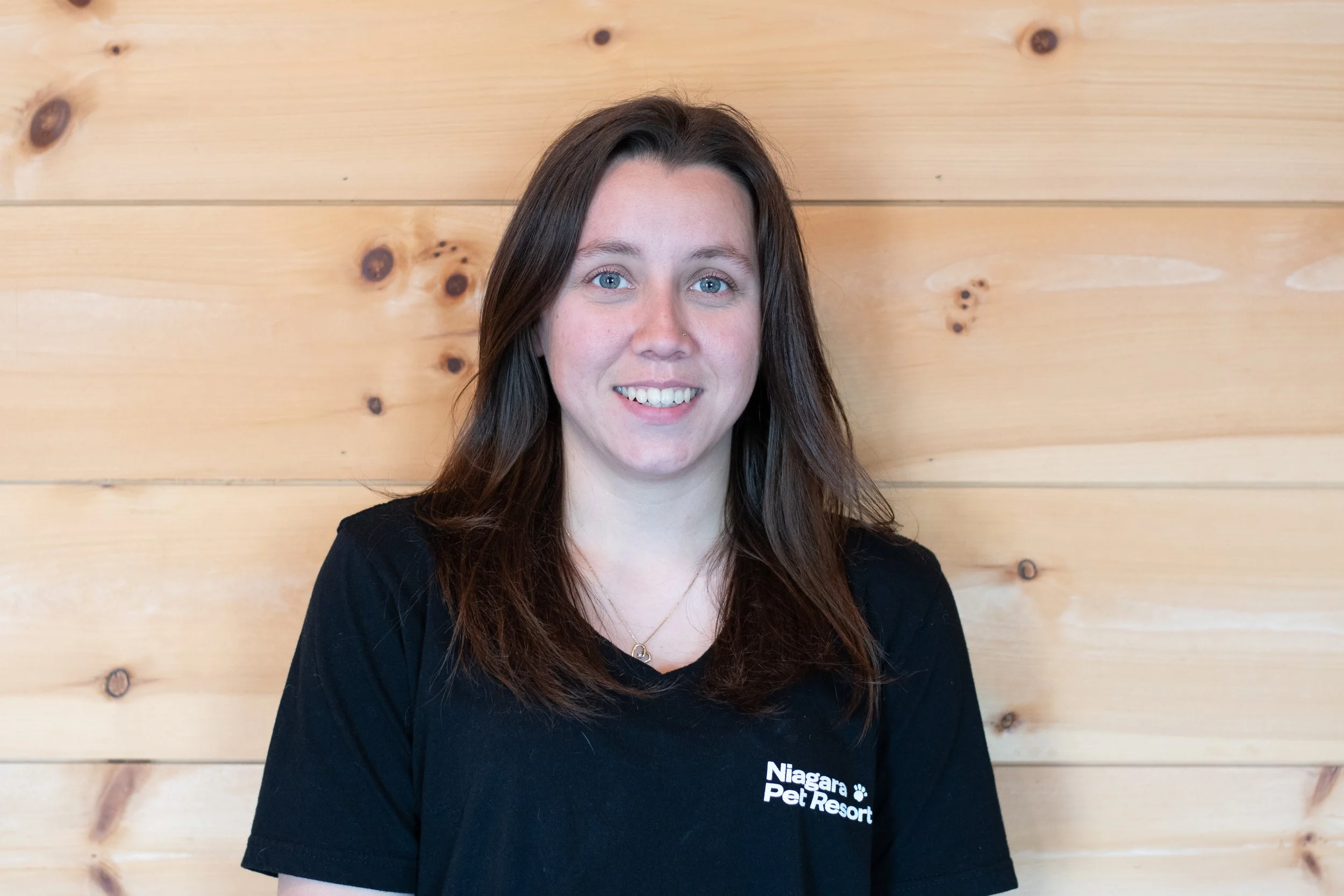 A woman with long brown hair and blue eyes smiling, wearing a black T-shirt with white text, standing in front of a wooden wall.