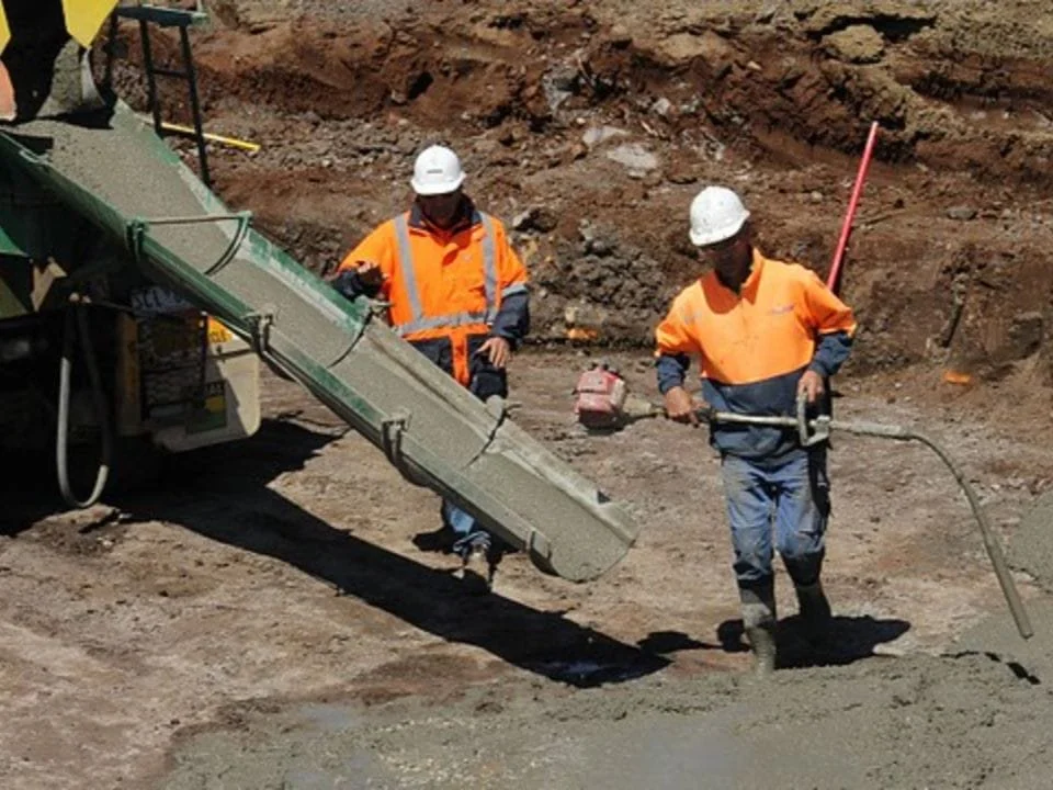 Two construction workers in orange safety shirts and white helmets working on a concrete pouring site with a concrete mixer truck.
