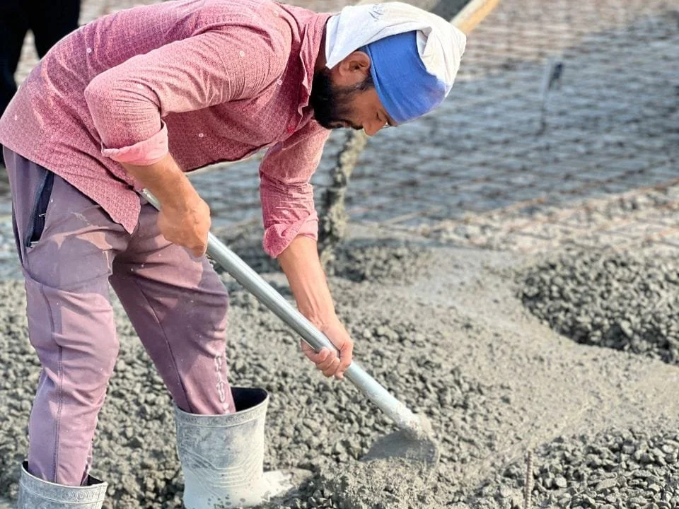 A person wearing a pink shirt, purple pants, and rubber boots is shoveling wet concrete on a construction site.