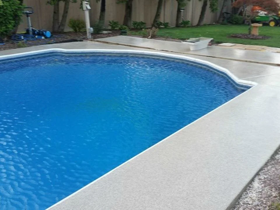 A backyard swimming pool with clear blue water, surrounded by a concrete deck, with a wooden fence and some plants in the background.