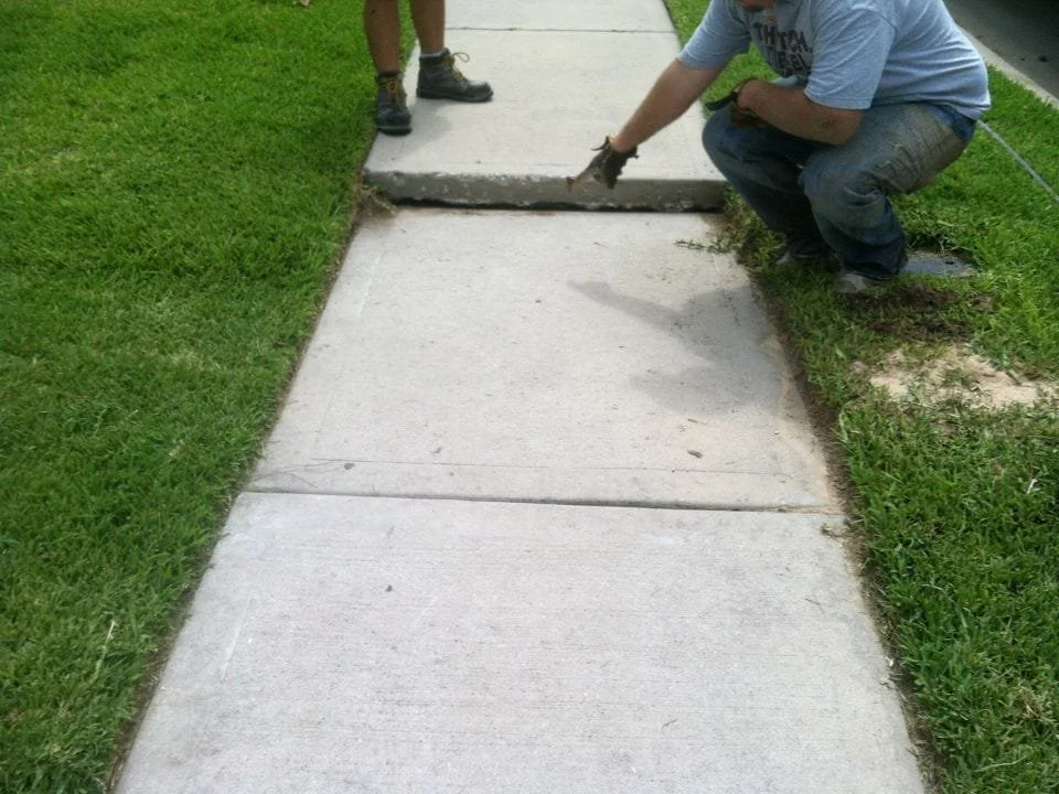 Man kneeling down and working on a concrete sidewalk with another person standing nearby, grass on both sides.