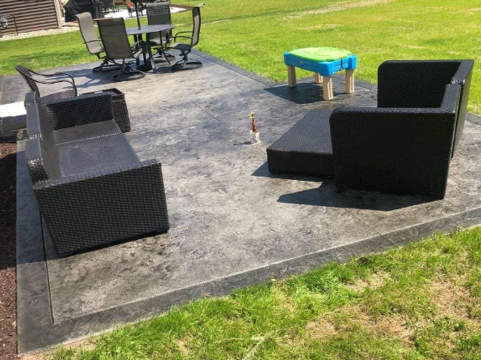 Outdoor patio area with black wicker chairs, a black wicker lounge chair with a cushion, a table with six chairs, and a small colorful plastic sandbox, surrounded by grass.