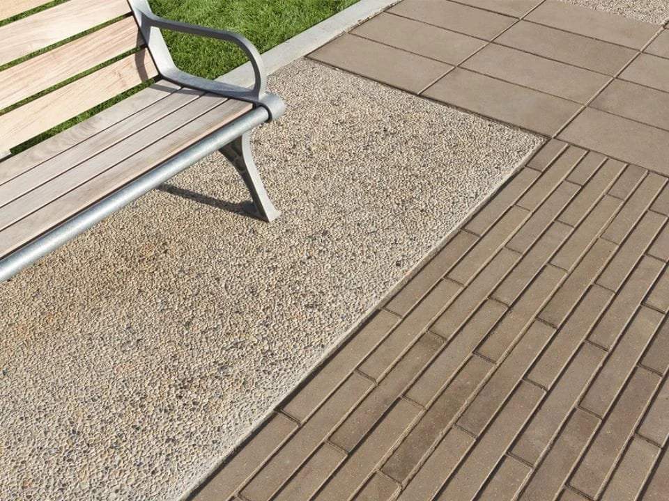 Empty park bench on a sidewalk with textured concrete and brick paving, near a grass area.