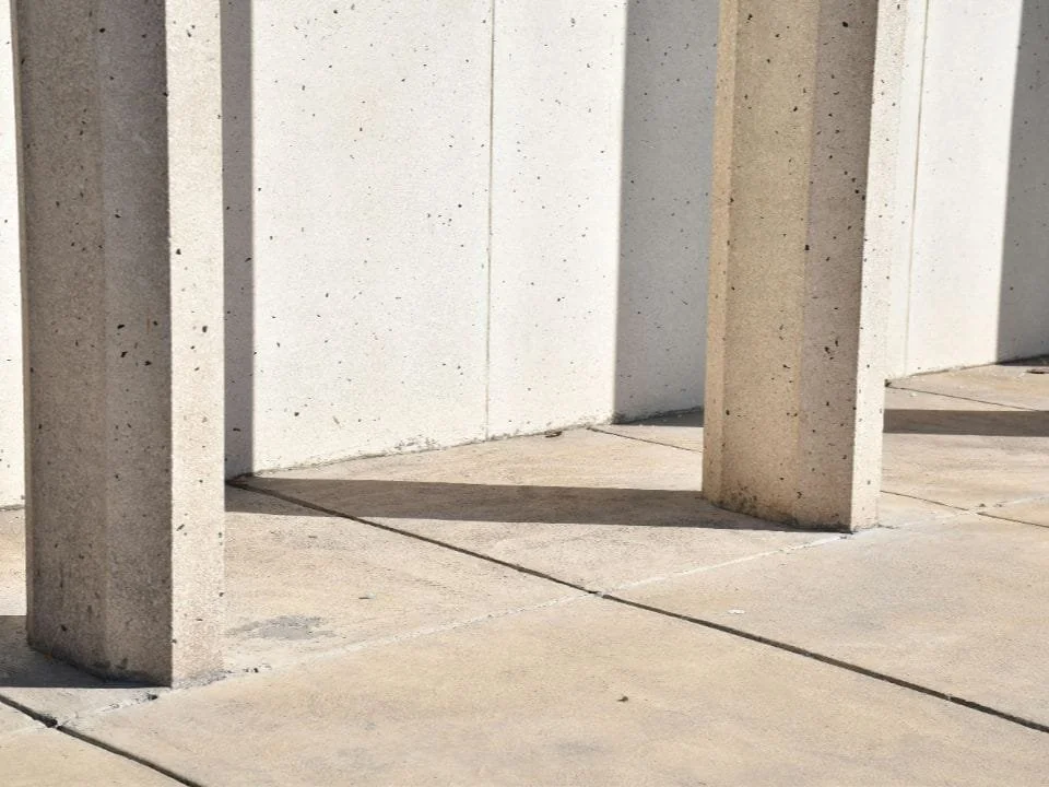 Concrete columns casting shadows on a concrete sidewalk outside a building.