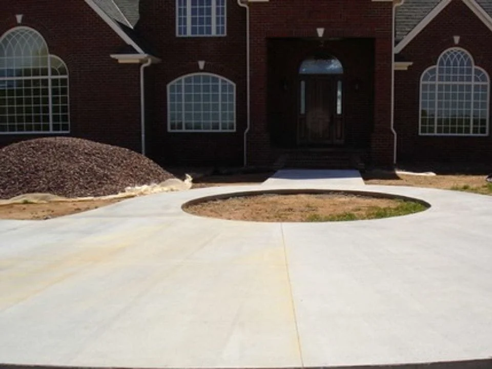 Newly paved concrete sidewalk with a circular bed of soil in front of a brick house with large arched windows and a dark wooden door.