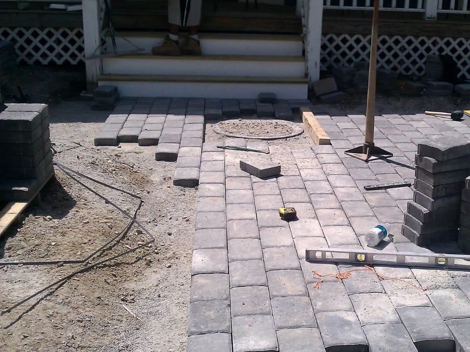 Paving stones are being laid to create a new walkway in front of a house, with construction tools and materials visible.