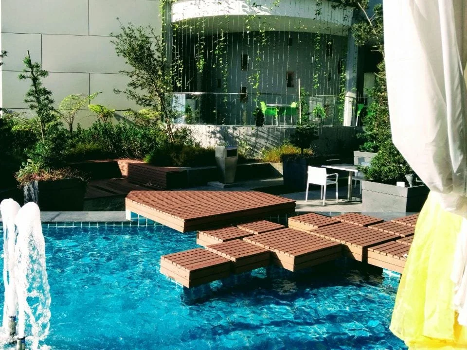 Poolside area with a wooden deck, greenery, and outdoor furniture, adjacent to a modern building with glass windows.