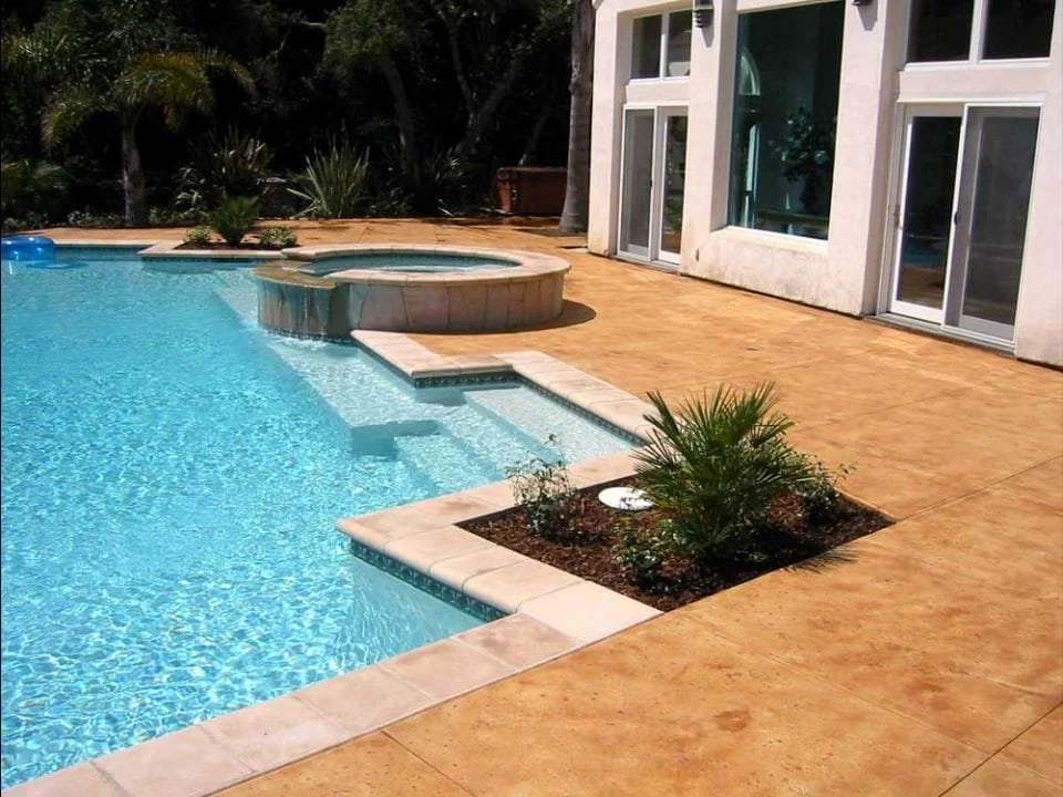 A backyard swimming pool area with a hot tub, a small plant bed, and a house with large glass doors.