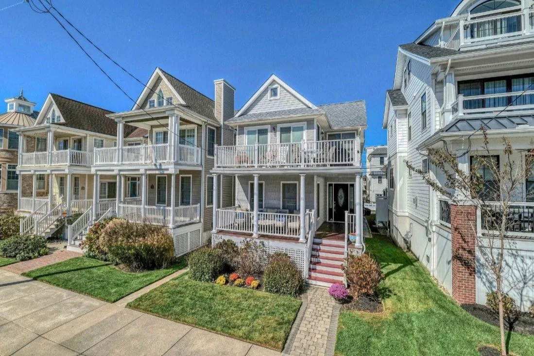 A row of residential houses with front porches, lawns, and landscaping in a suburban neighborhood under a clear blue sky.