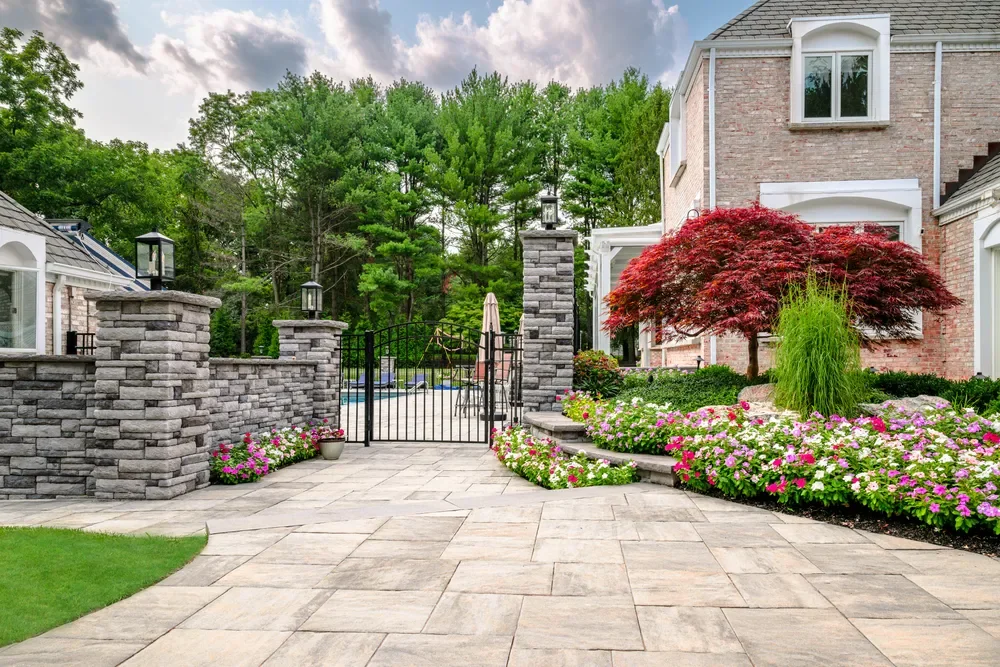 View of a landscaped backyard with stone patio, stone privacy wall with lanterns, black gate, colorful flowers, a red-leafed tree, and a brick house in the background, surrounded by tall green trees under a cloudy sky.