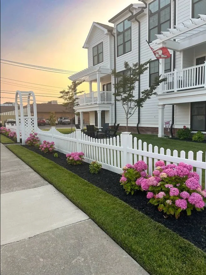 A white porch with garden furniture, a small tree, and a white picket fence with pink hydrangea flowers in a residential neighborhood at sunset.