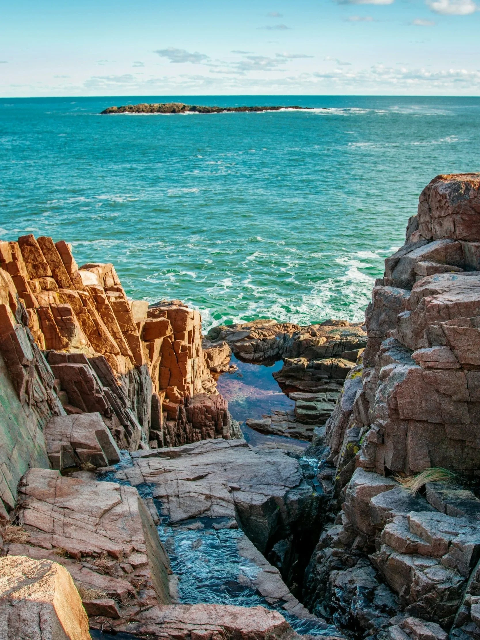 Rocky coastline with turquoise ocean water, a small island in the distance under a partly cloudy sky.